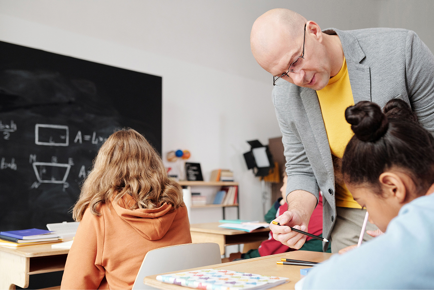 Teacher in glasses and gray blazer helps students with work in a classroom with chalkboard.