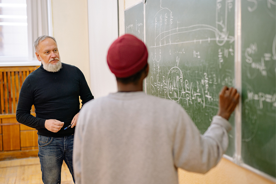 Teacher watching student in red hat write math equations on a chalkboard in a classroom.