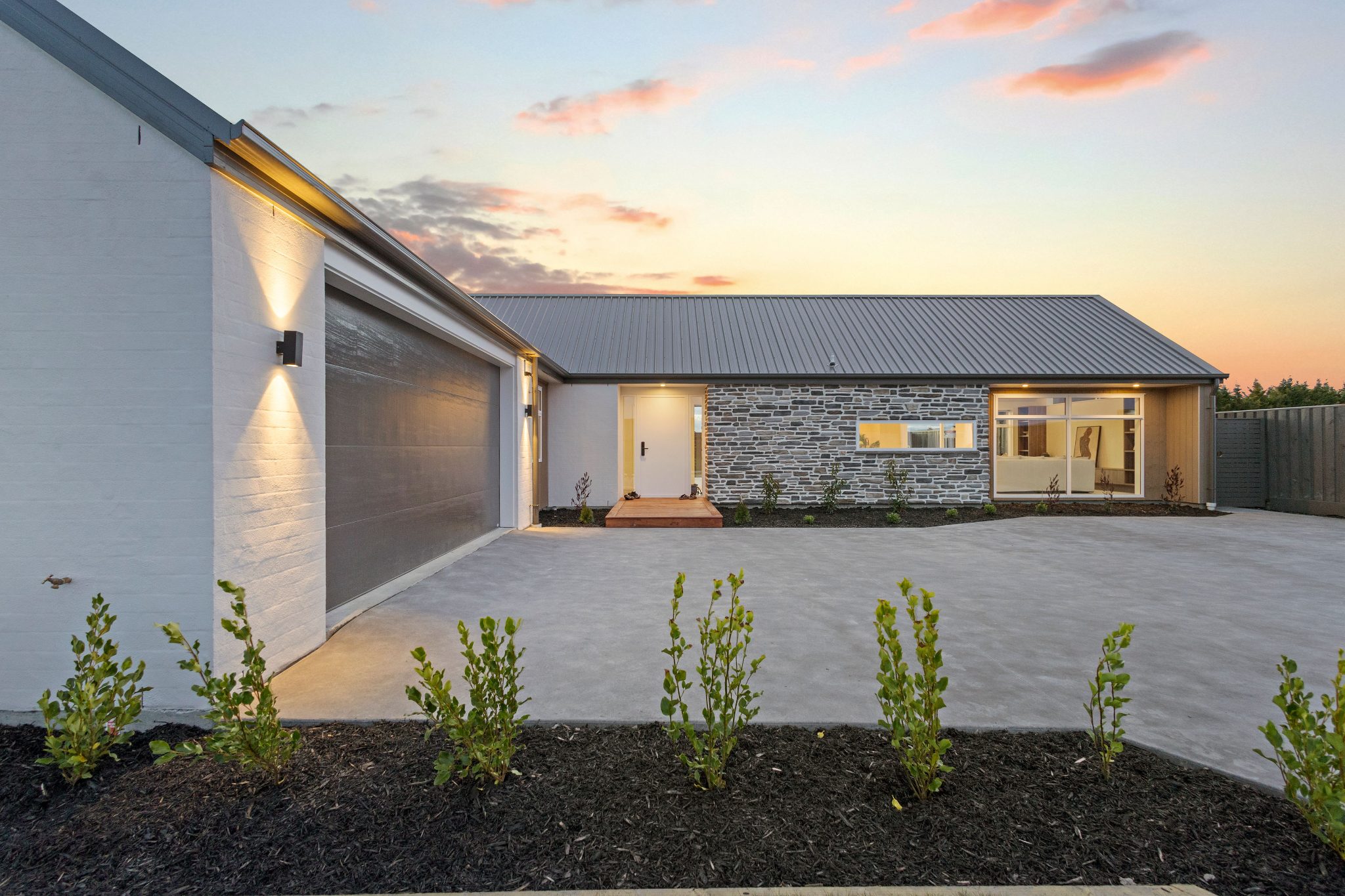 Modern single-story house with stone facade, large garage door, and a concrete driveway at sunset.