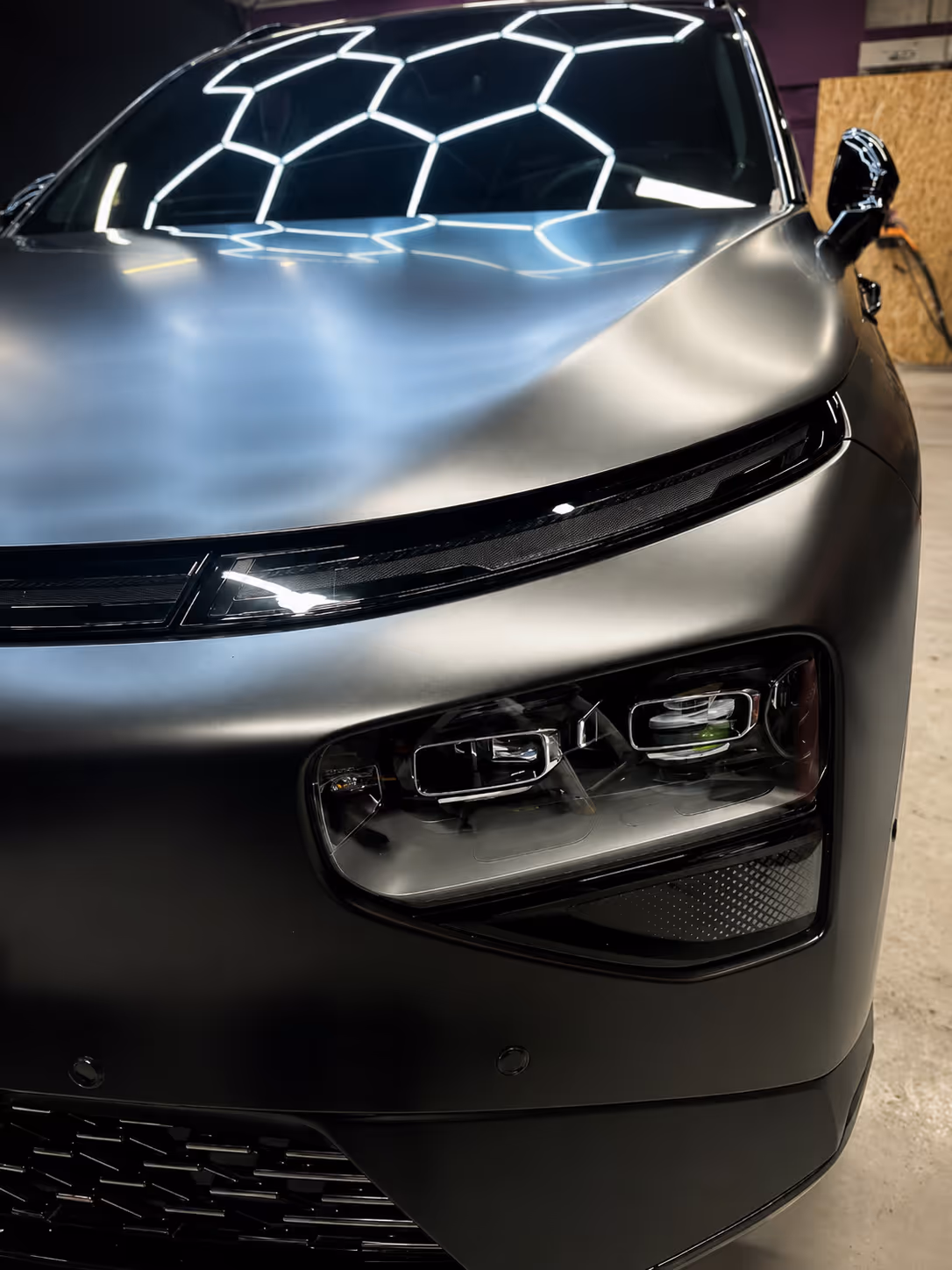 Close-up of a sleek matte black car front with modern headlights and geometric ceiling reflection.