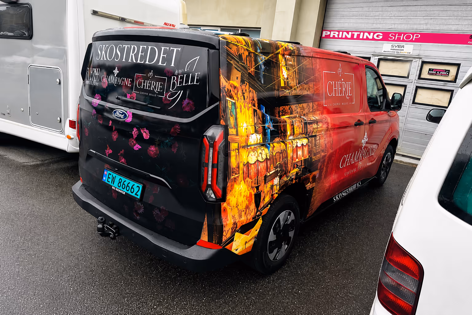 Black and red Ford van with floral and cityscape graphics parked next to white vans on wet pavement.