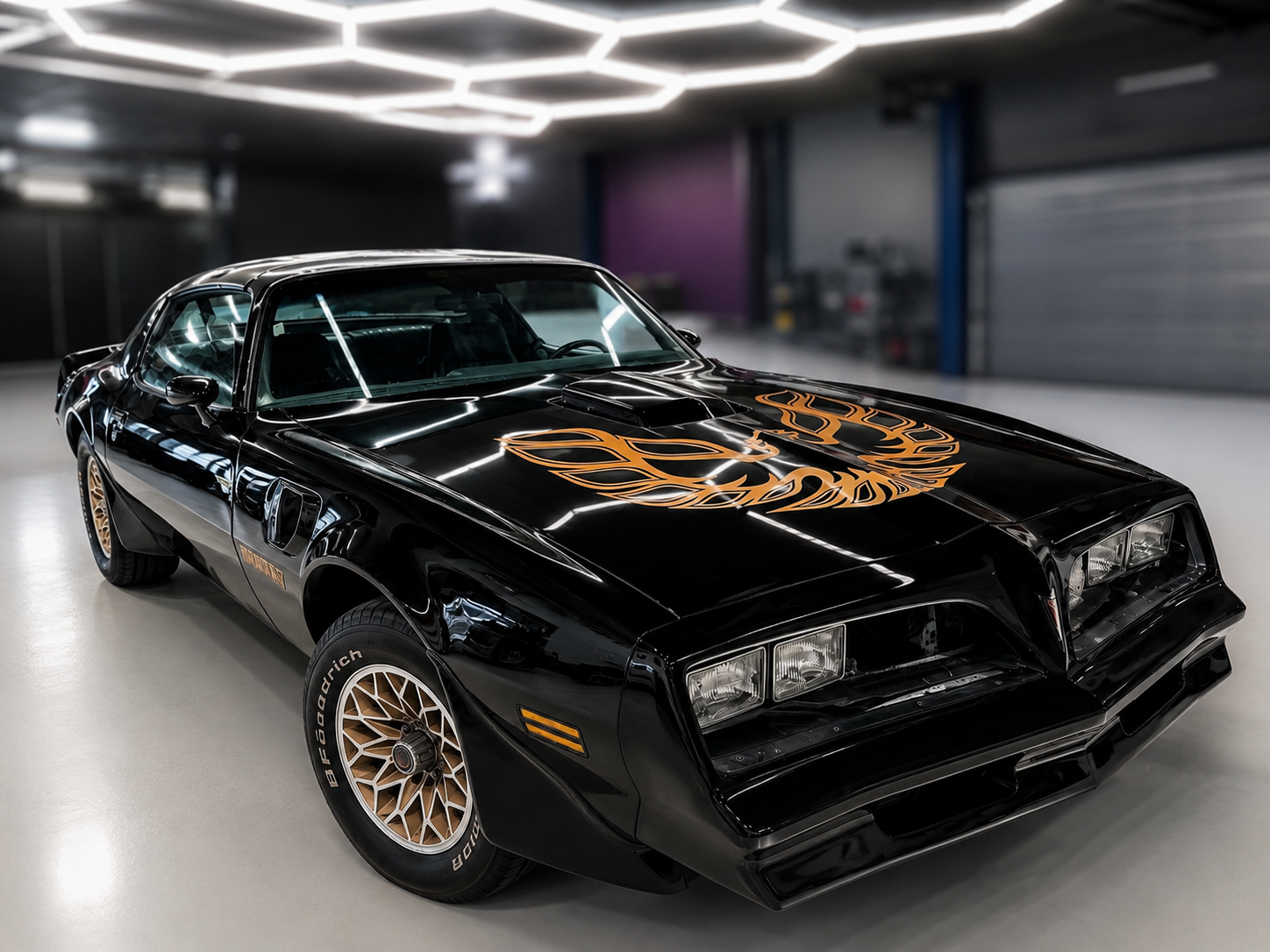 Black classic Pontiac Firebird Trans Am with gold phoenix decal on hood and gold rims, in a garage.