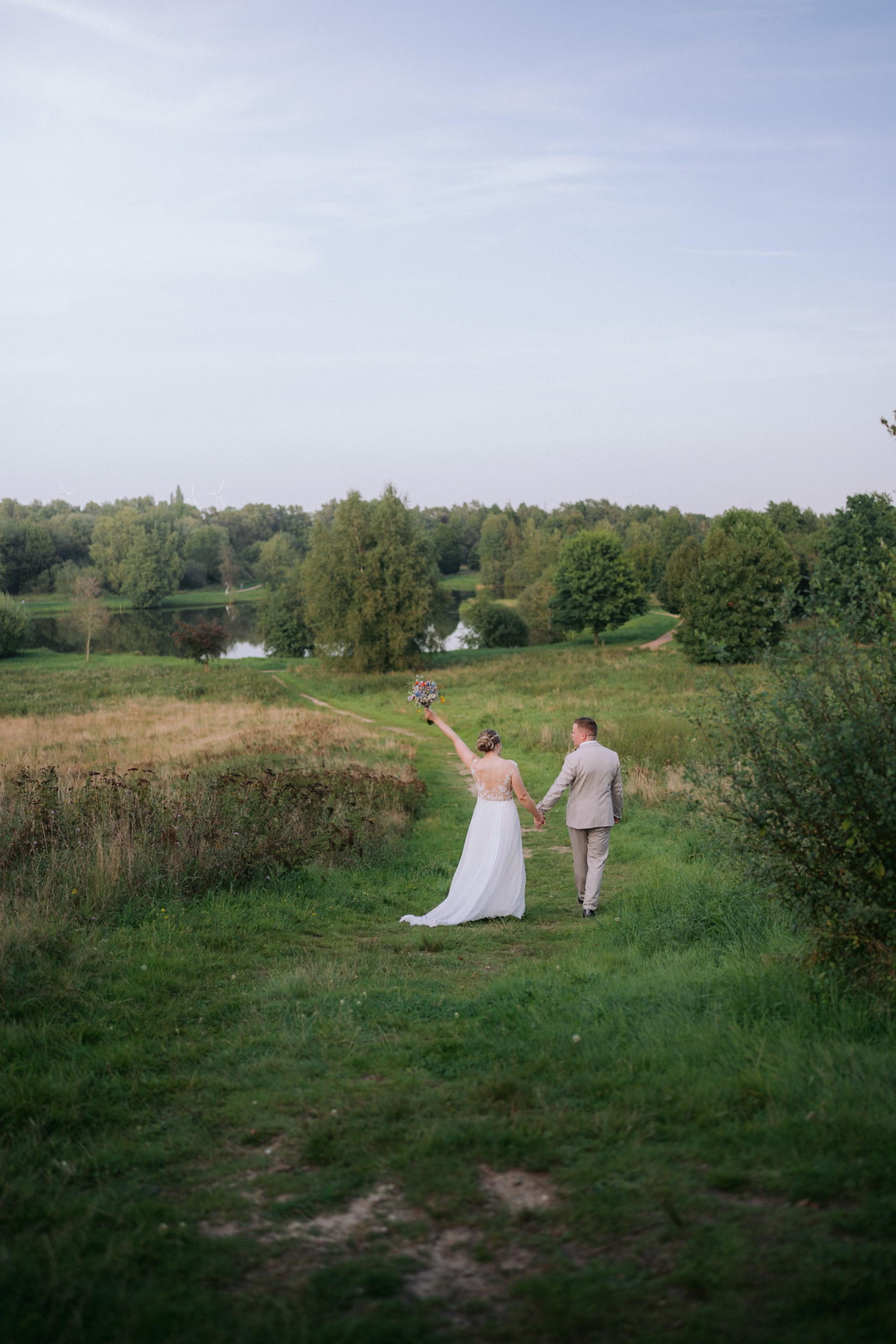 Fenja und Erik gehen Hand in Hand über einen Feldweg, sie hält den Brautstrauß hoch, dahinter ein See