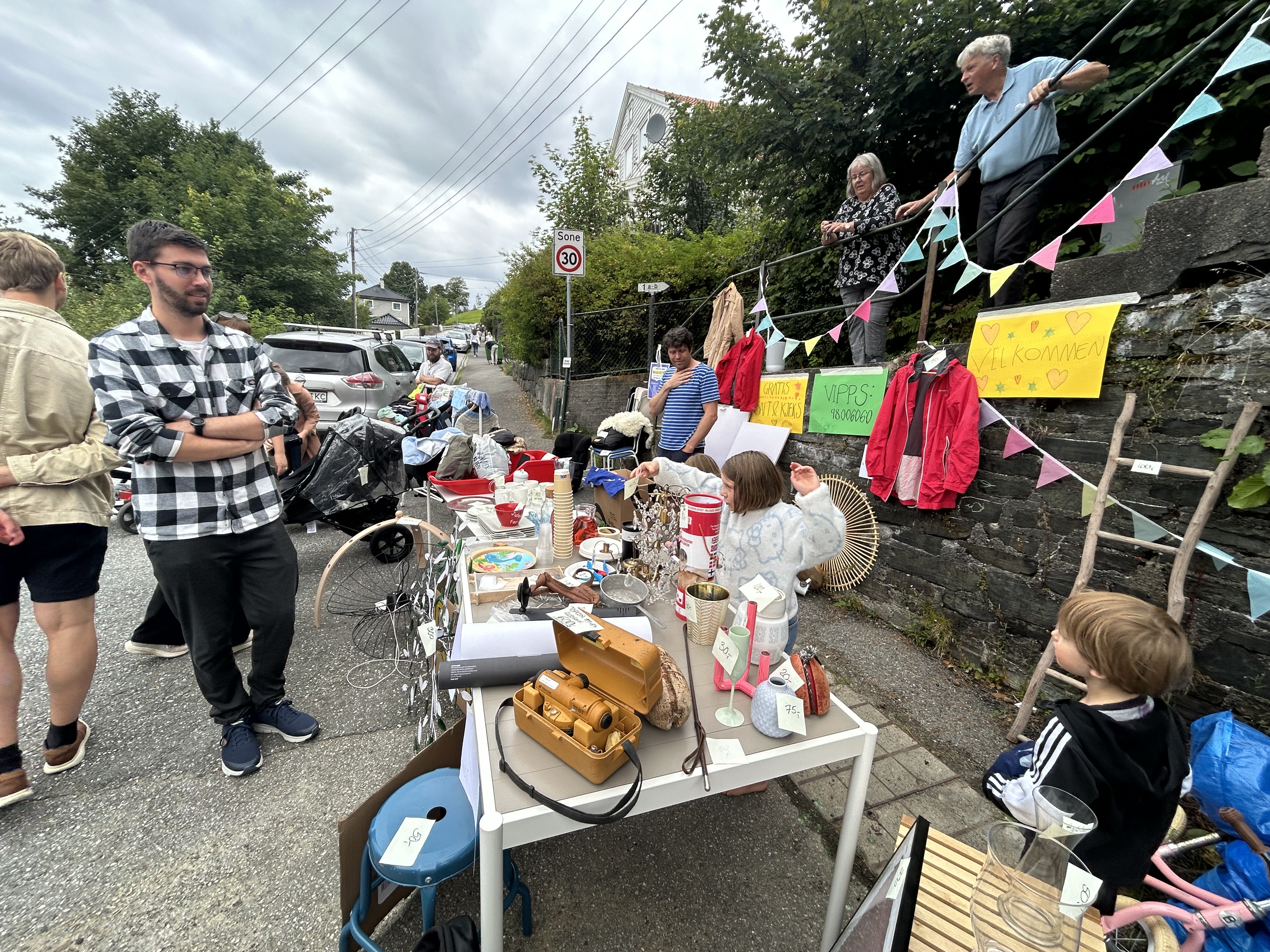 Folk samlet på gateauksjon med bord dekket med diverse gjenstander til salg under overveiende skyet himmel.