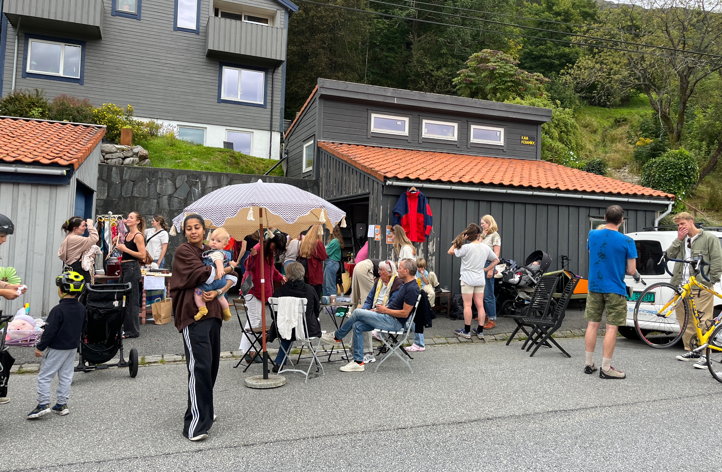 Folk samles utendørs ved et lite bygg med paraply, stoler og sykler i et boligområde.