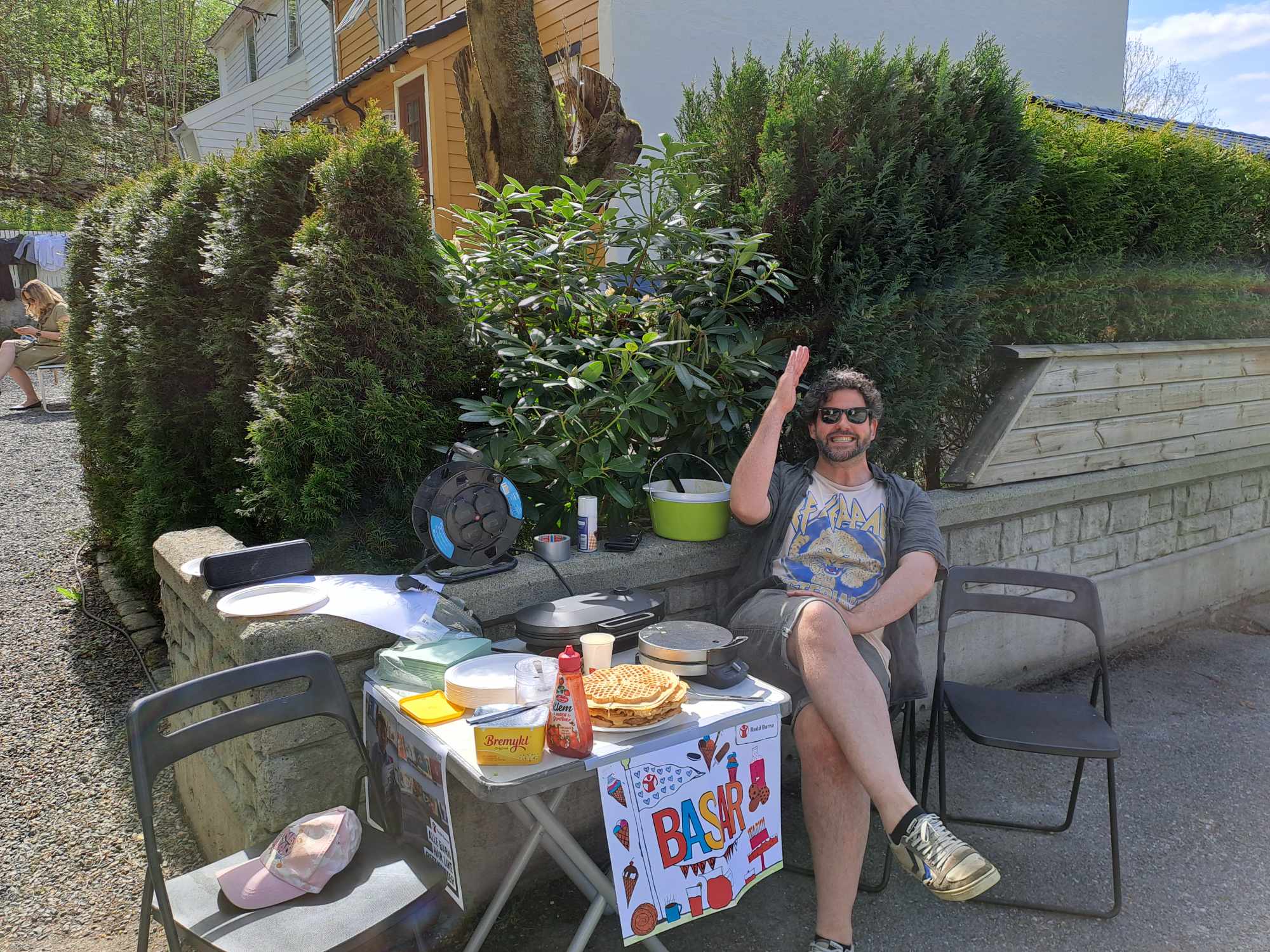 Mann med solbriller sitter ved bord med vafler og basar-skilt utendørs, smilende og vinkende.