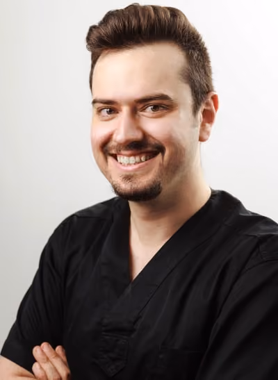 Smiling man with short hair and goatee wearing black scrubs with folded arms on light background.