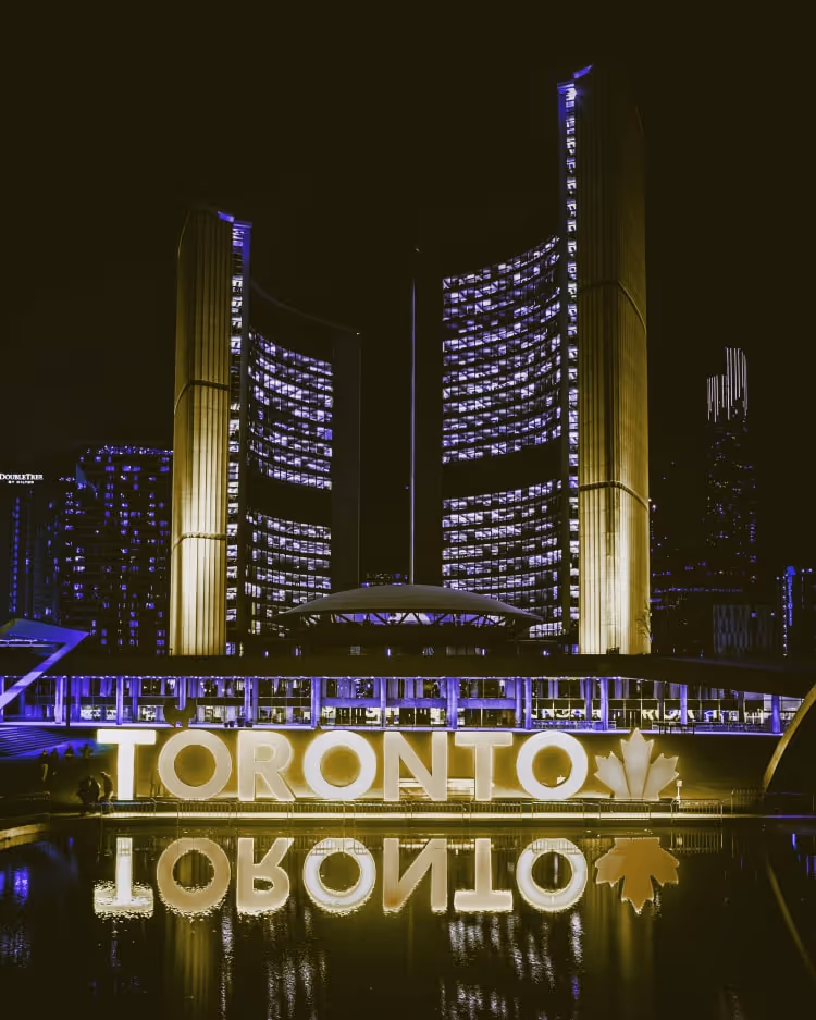 Night view of Toronto's illuminated city hall with glowing TORONTO sign reflecting on water.