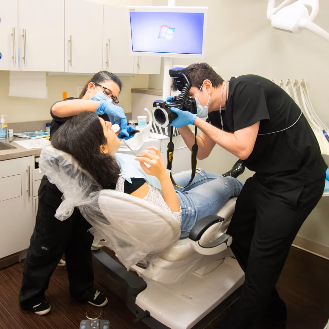 Dentist and assistant wearing masks and gloves examine patient’s teeth using a camera in dental clinic.