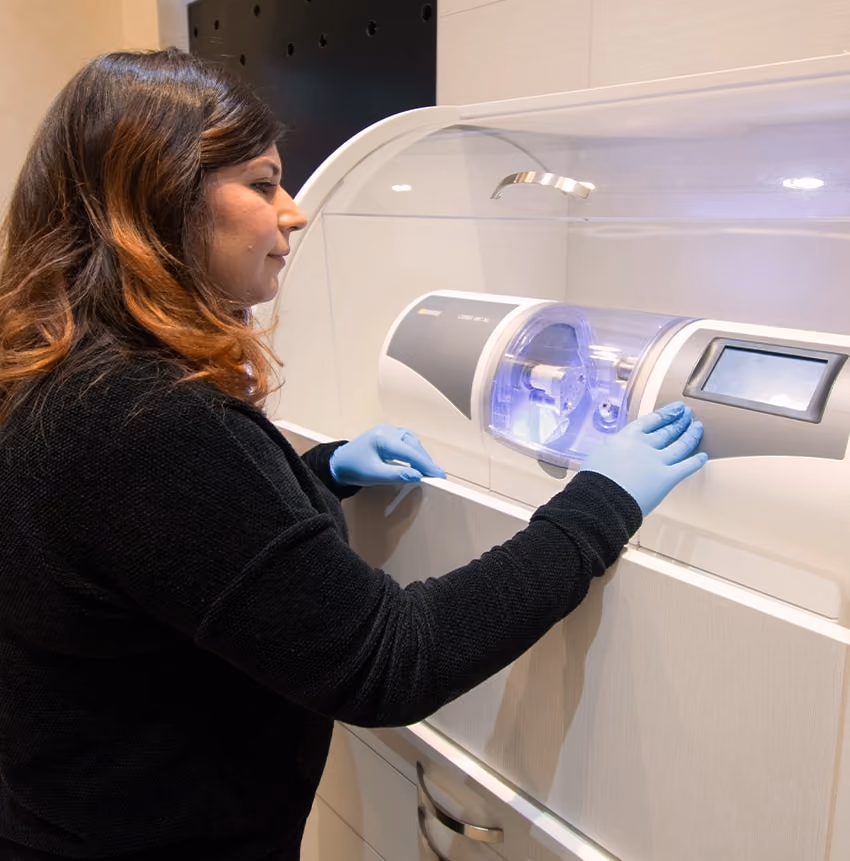 Woman wearing blue gloves operating a dental machine with a touchscreen in a clinical setting.
