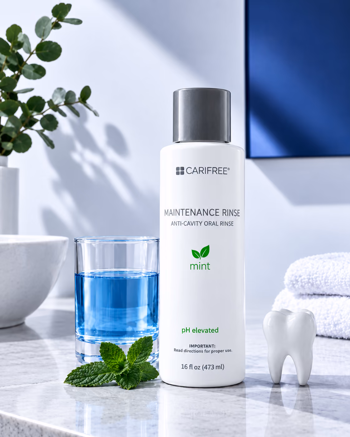 A bottle of Carifree Maintenance Rinse mint-flavored anti-cavity oral rinse on a marble countertop, next to a glass with blue liquid, mint leaves, a large tooth model, and white towels in the background.