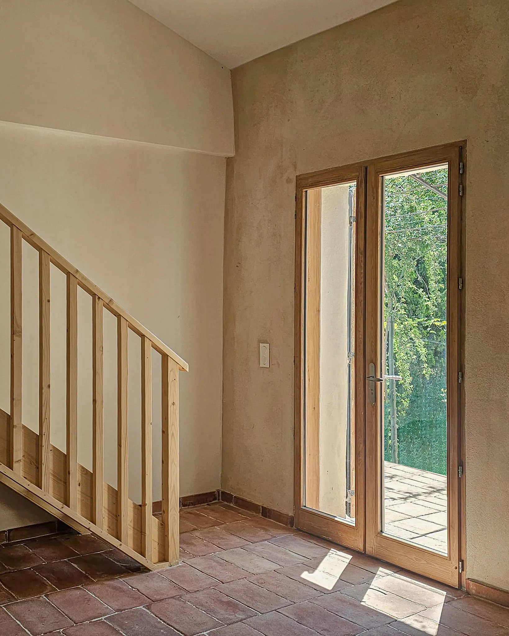 Empty room with tiled floor, wooden staircase railing, and glass door opening to a sunny outdoor area.