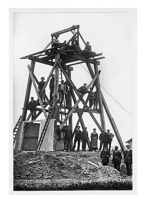 A group of men and one woman posing on and around a tall wooden construction framework coal mining.