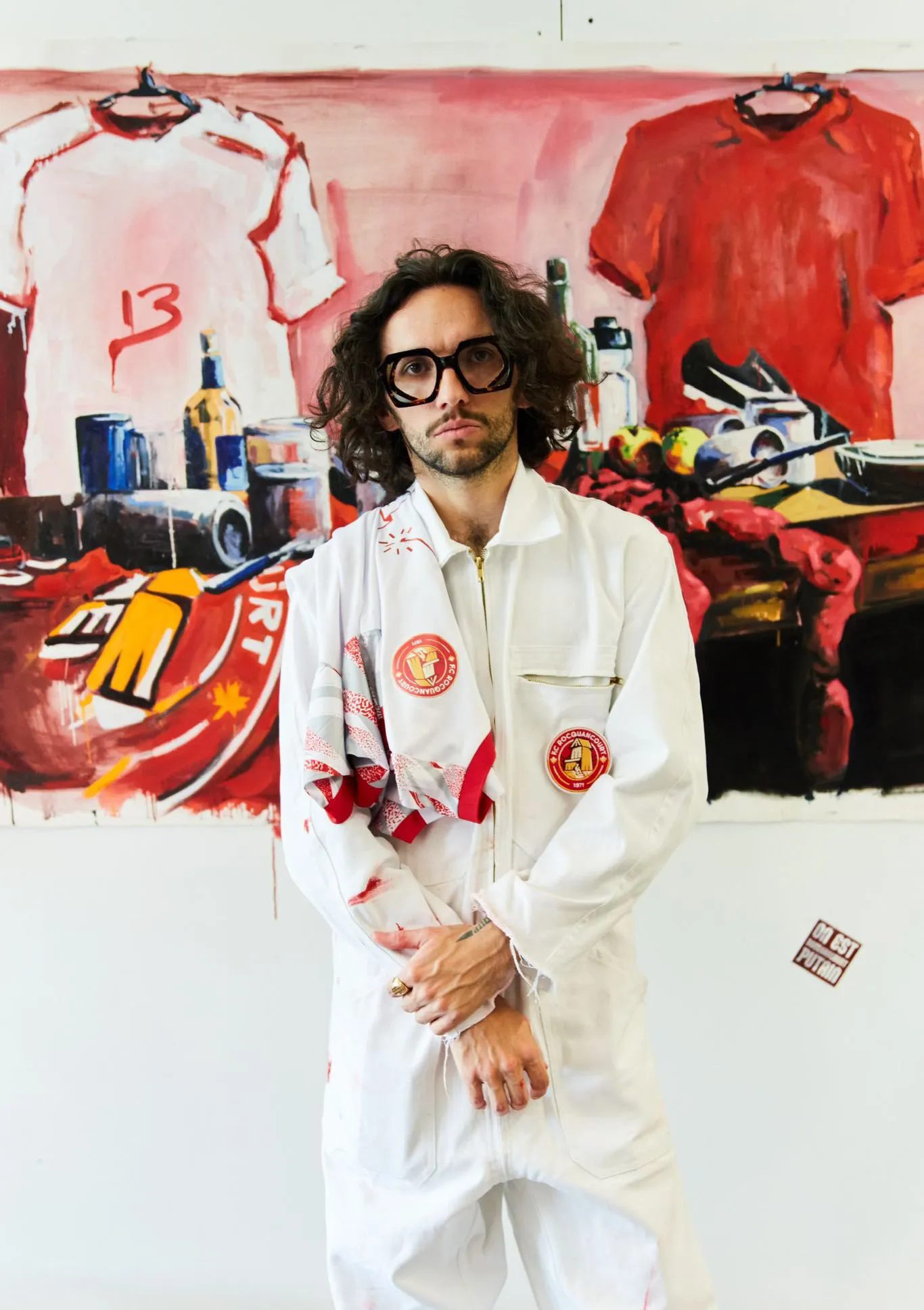 Man with glasses in white jumpsuit stands in front of a colorful abstract painting with shirts and bottles.