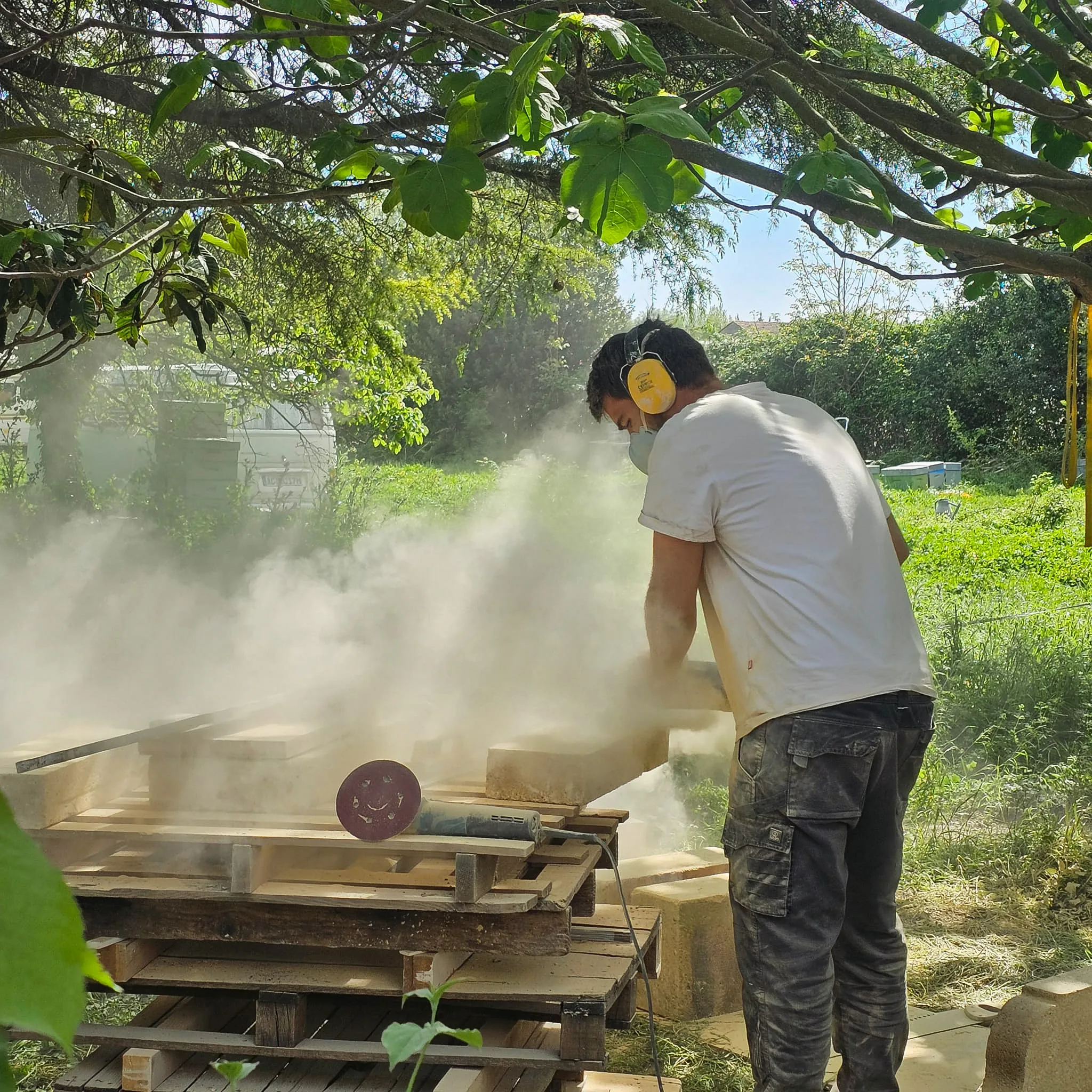 Man wearing ear protection and mask cutting stone outdoors with dust in the air under tree branches.