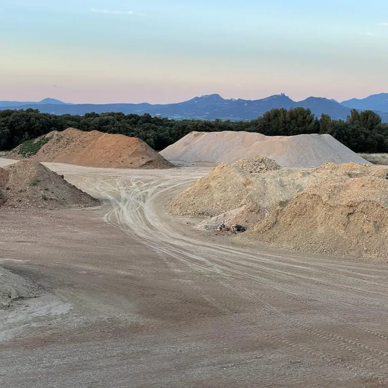 Dirt and gravel piles on a cleared area with tire tracks, trees in background, and mountains at sunset.