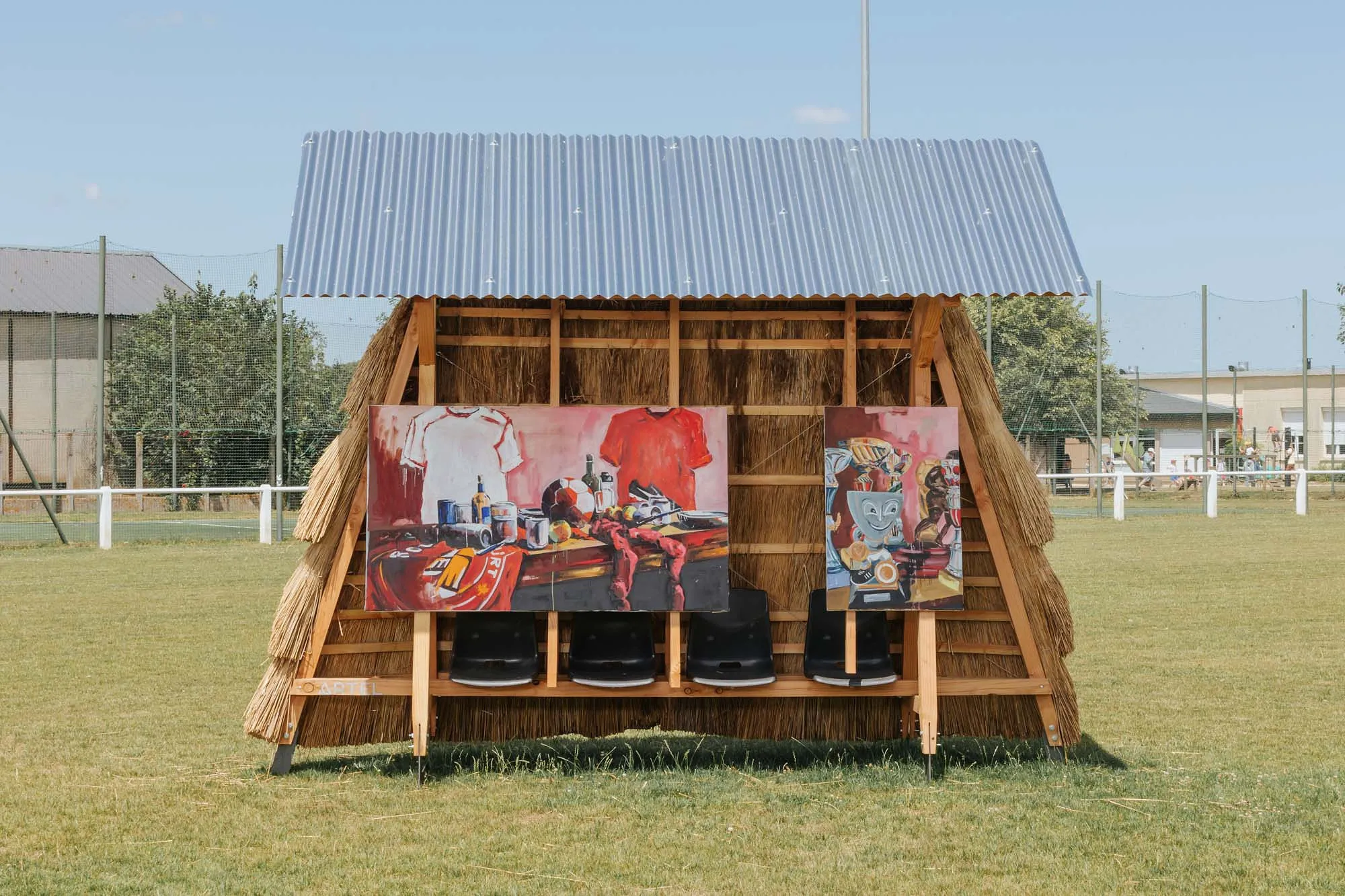 Thatched wooden shelter with two colorful paintings displayed on seats in a grassy field under a blue sky.