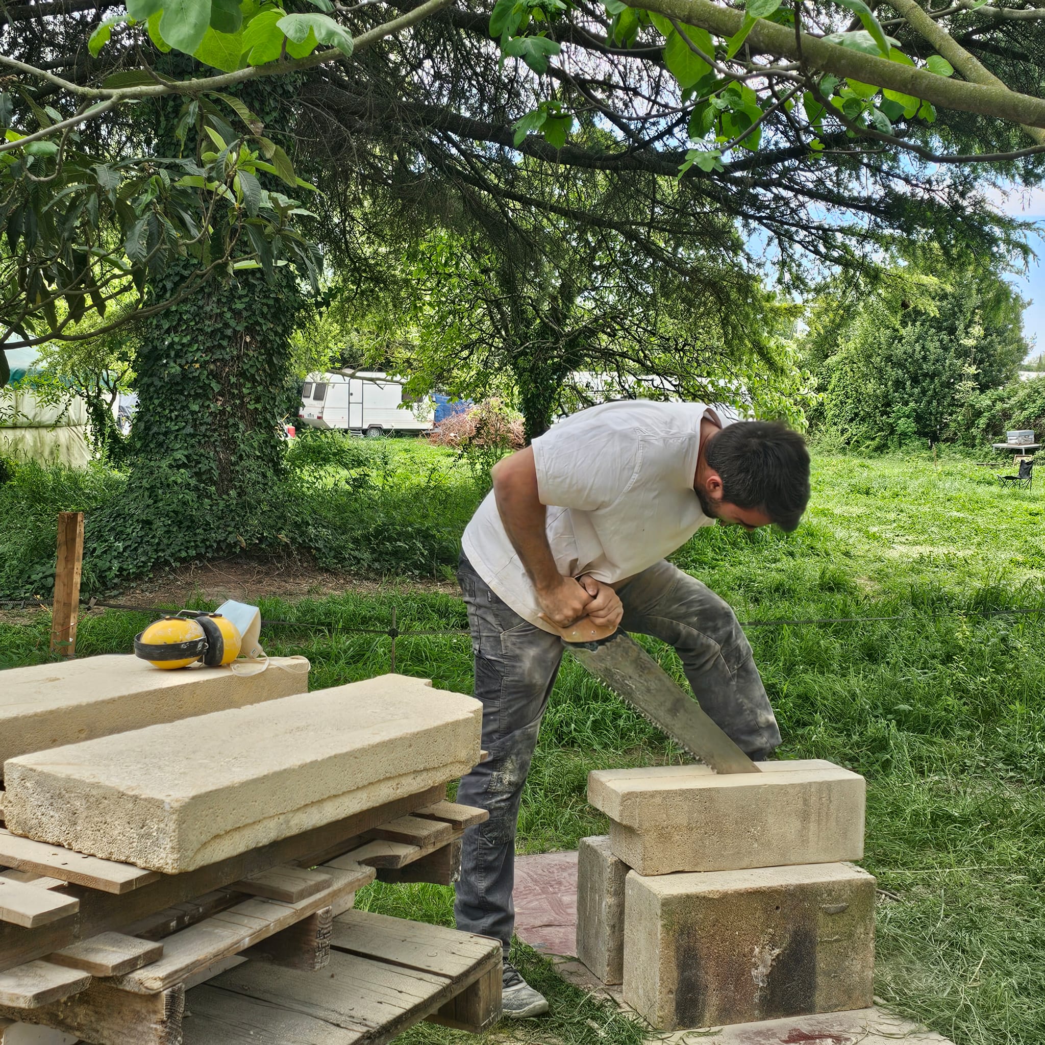 Man wearing white shirt and dusty pants saws a stone block outdoors with greenery around.