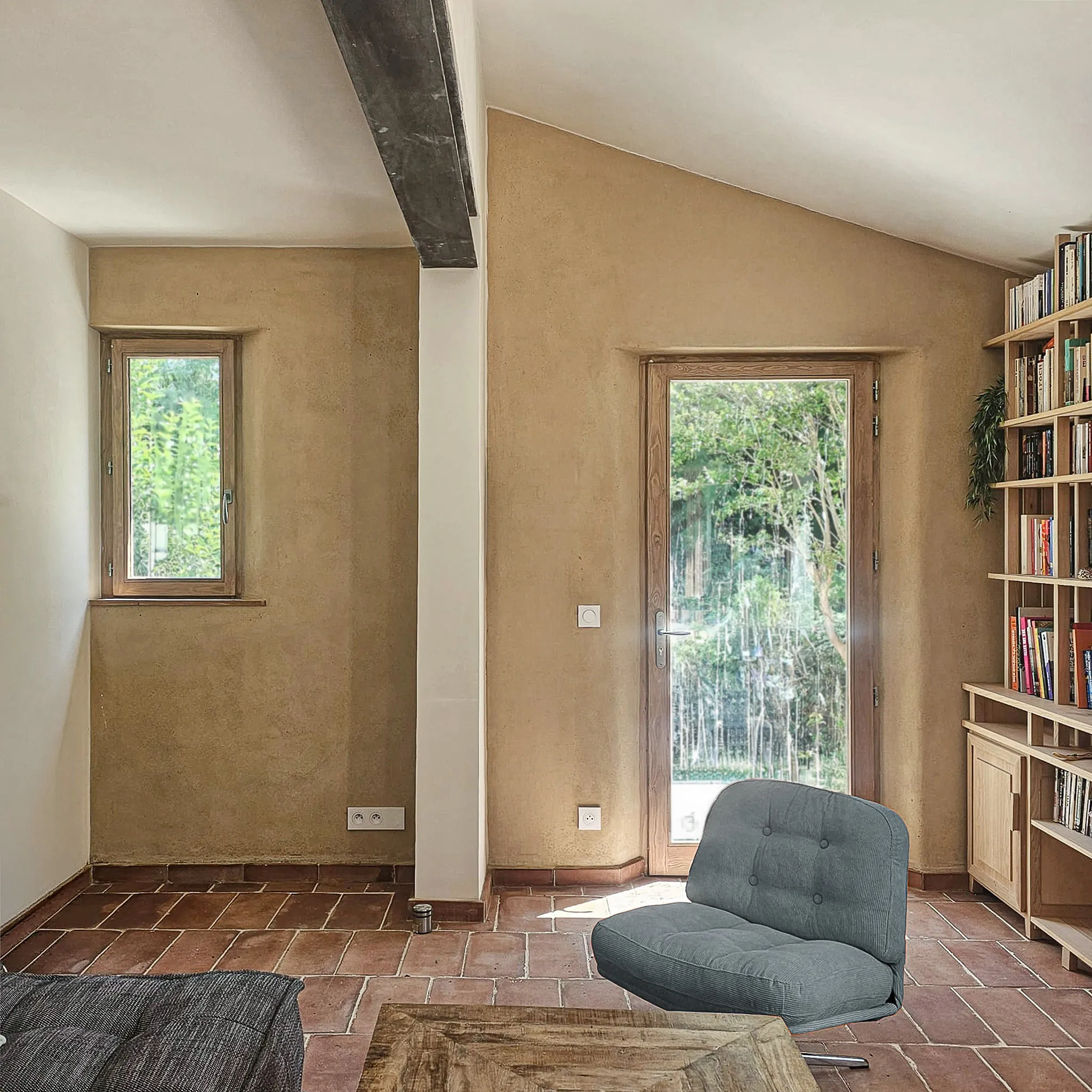 Room with terracotta floor, wooden bookshelf, glass door, window, gray cushioned chair, and wooden table.