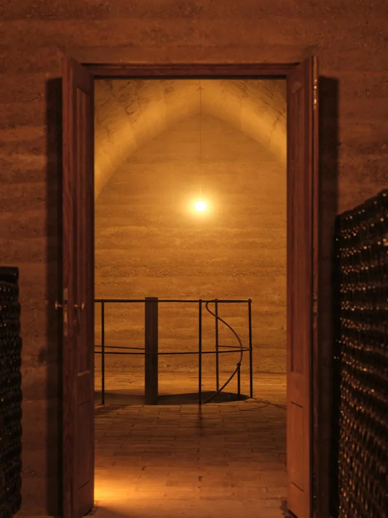 Open wooden doors reveal a warmly lit stone cellar with an arched ceiling and a metal railing inside.