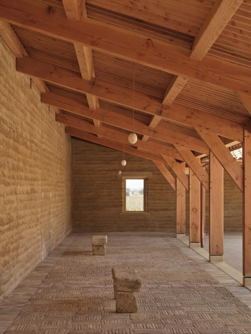 Indoor space with wooden beams and pillars, textured floor, site concrete walls, and rectangular window.