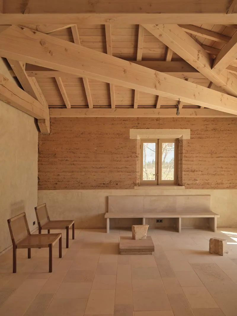 Minimalist room with wooden ceiling beams, two wooden chairs, a bench, and stone blocks on tiled floor.