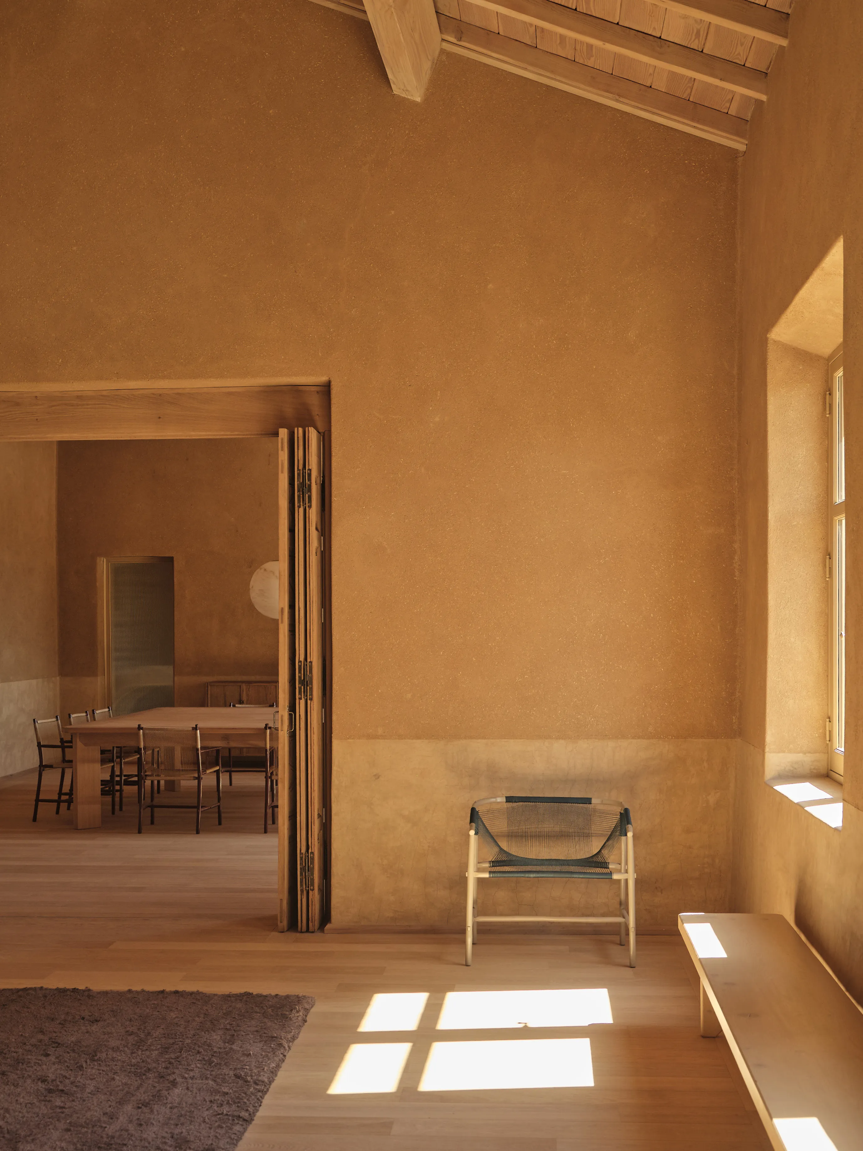 Minimalist room with beige walls, a woven chair, wooden bench, and sunlight through a window.