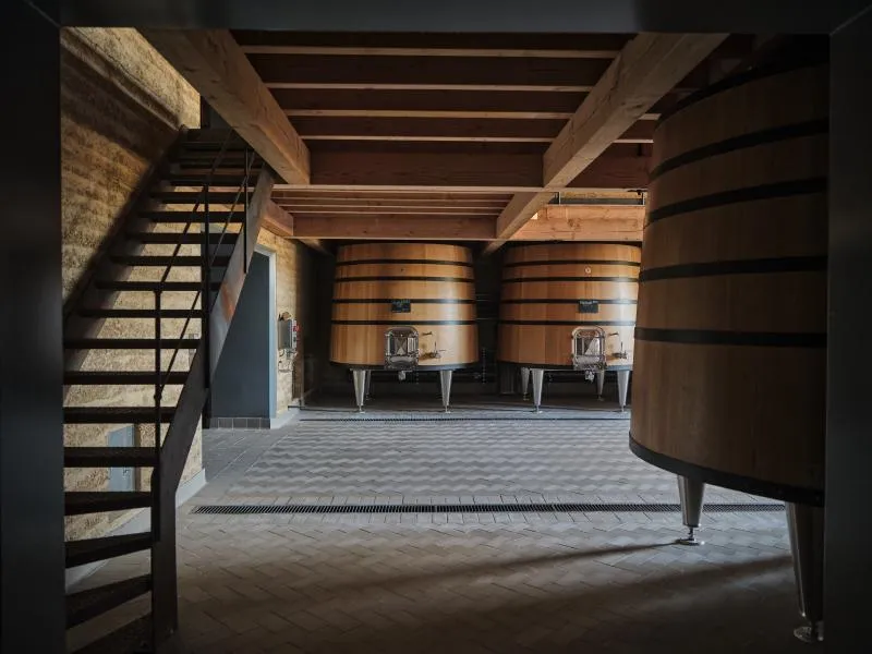 Interior of a cellar with large wooden fermentation tanks and a metal staircase to the left.