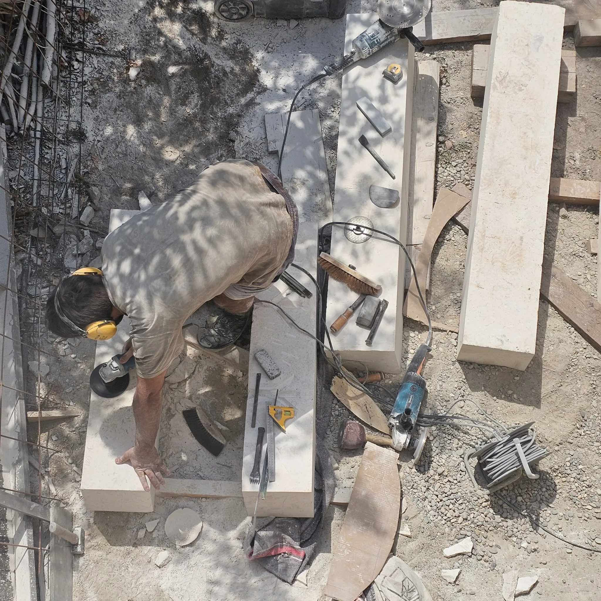 Worker wearing ear protection polishing large stone slabs with power tools on dusty ground.