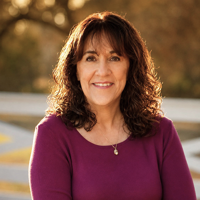 A portrait of Darleen in an Autumn outdoor setting wearing a burgundy sweater with her brown hair down, exhibiting a soft, warm smile.