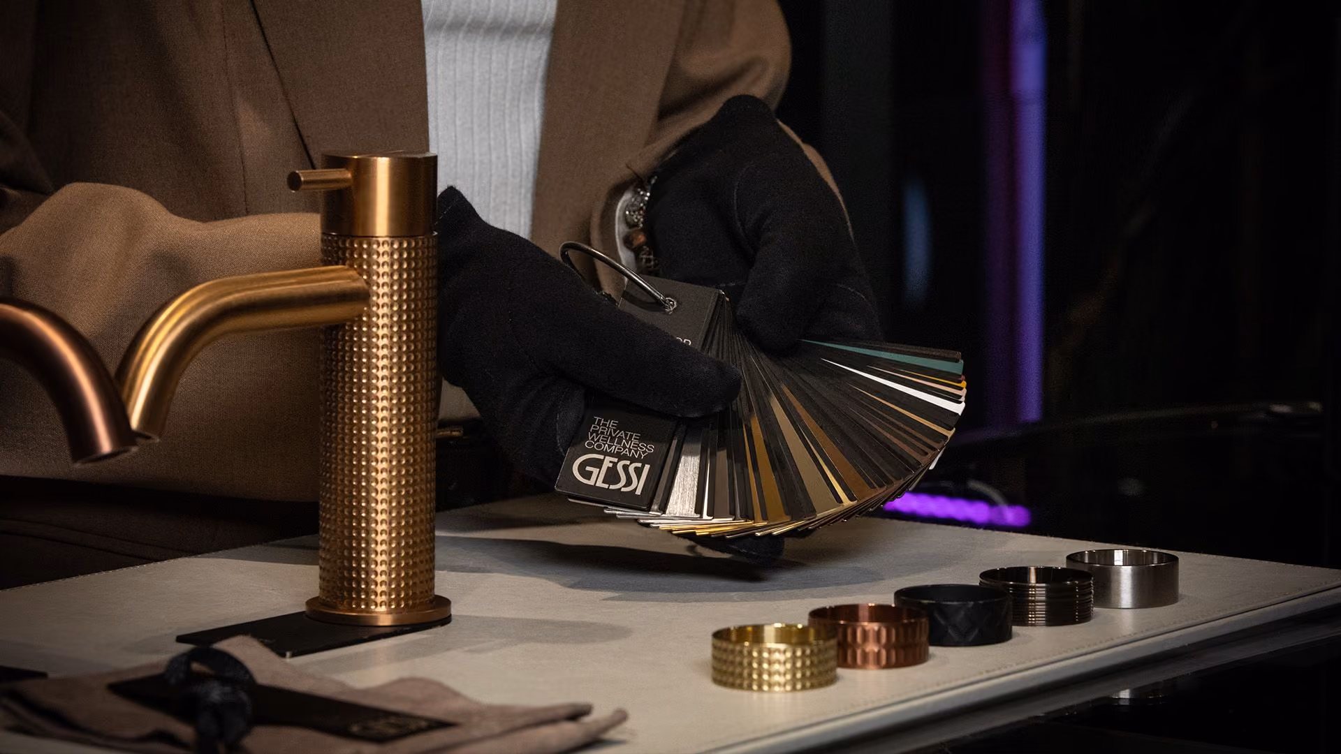 Person wearing black gloves holding a Gessi color finish sample fan displaying various dark and metallic shades next to a gold textured faucet and several matching circular metal rings on a table.