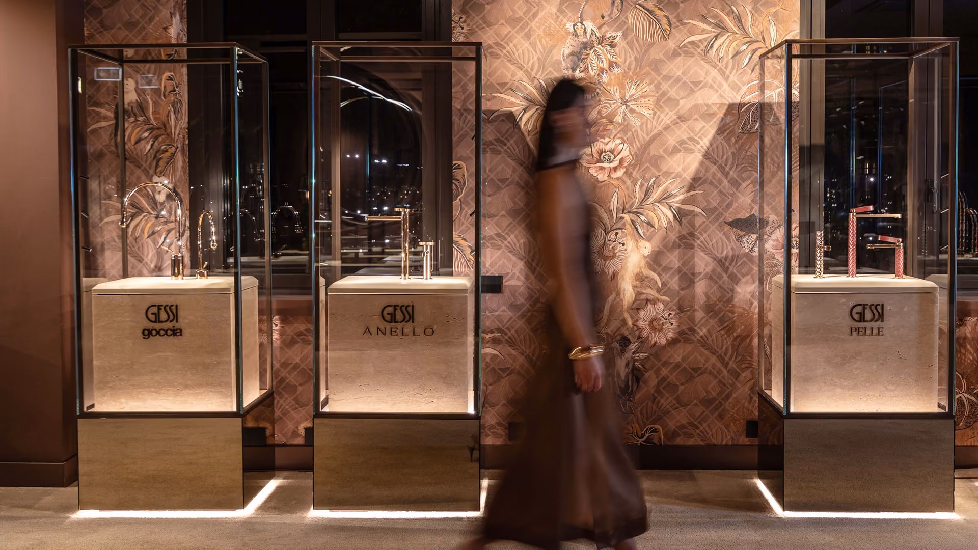 Three glass display cases each showcasing a different Gessi faucet model named Goccià, Anello, and Pelle, against a decorative wall with floral and animal motifs, with a woman walking past in a brown dress.