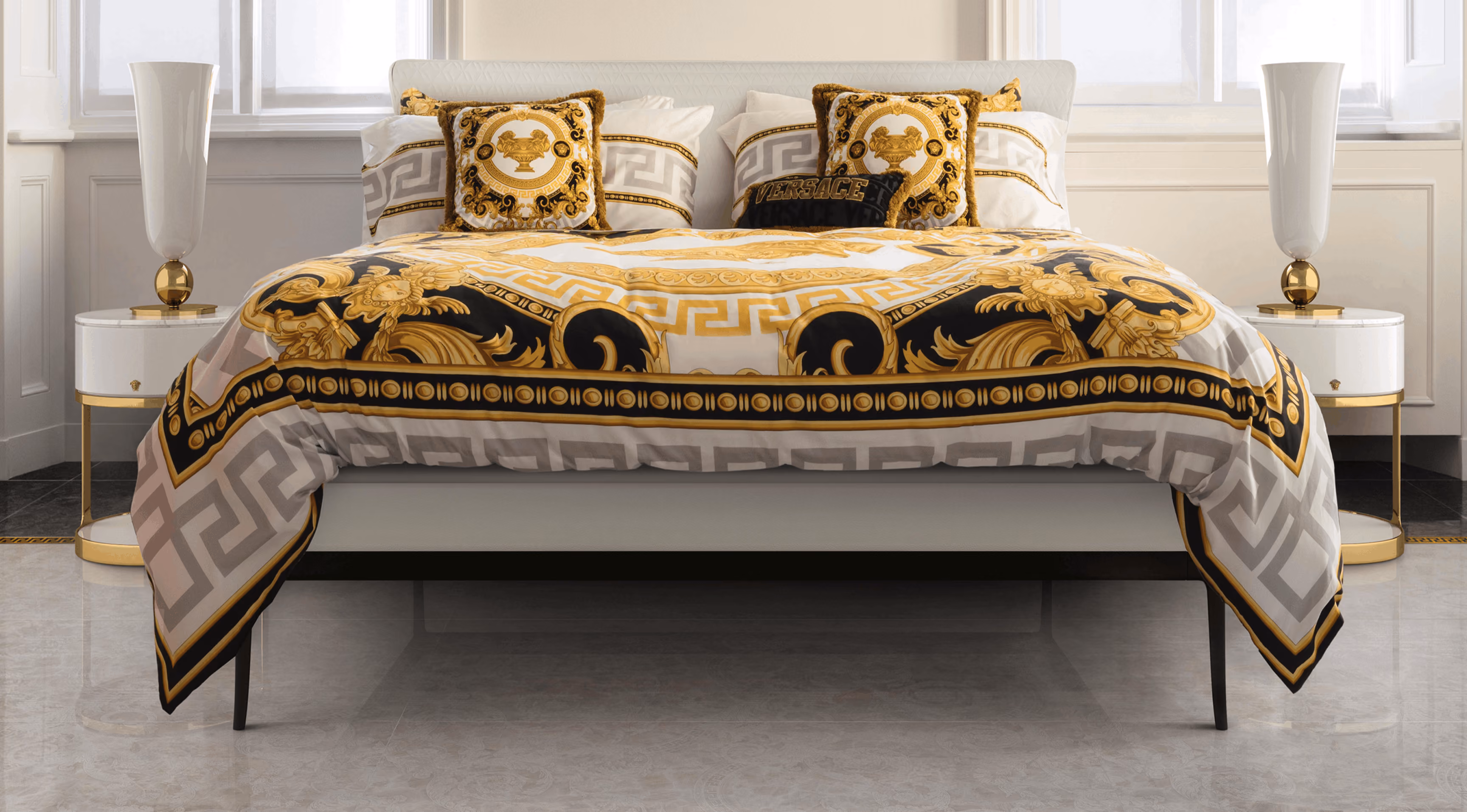 Luxurious bed with white frame, decorated with ornate bedding and pillows featuring gold, black, and white Baroque-style patterns, flanked by two modern white lamps on round white and gold side tables in a bright, elegant bedroom.