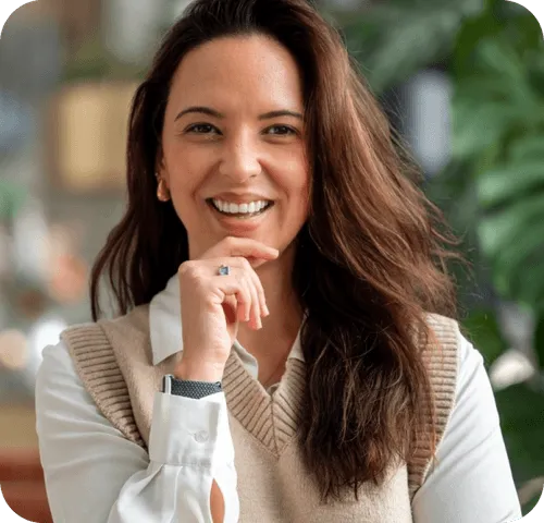 Smiling woman with long brown hair wearing a beige sweater vest and white blouse, posing indoors.