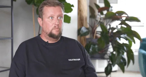 Man with beard wearing black Cole Buxton shirt sitting indoors with plants in the background.