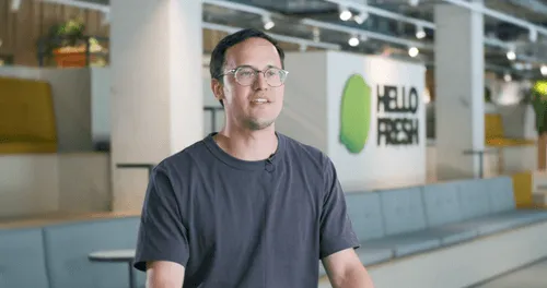 Man wearing glasses and a dark shirt speaking in a modern office with Hello Fresh logo in background.
