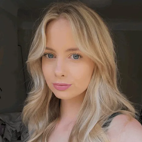 Blonde woman with wavy hair and blue eyes looking at the camera in soft light.