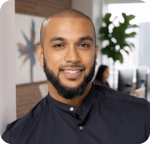 Smiling man with trimmed beard wearing a dark shirt in a modern office setting.
