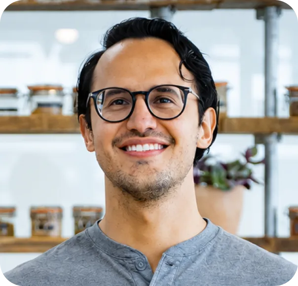 Smiling man with glasses wearing a gray shirt with shelves and jars blurred in the background.