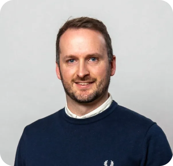 Smiling man with short brown hair and beard wearing a navy sweater with white shirt collar.