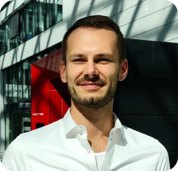 Man with short dark hair and beard wearing a white shirt, smiling, standing outdoors near modern building.