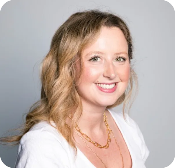 Smiling woman with wavy light brown hair wearing a white top and gold necklaces, gray background.