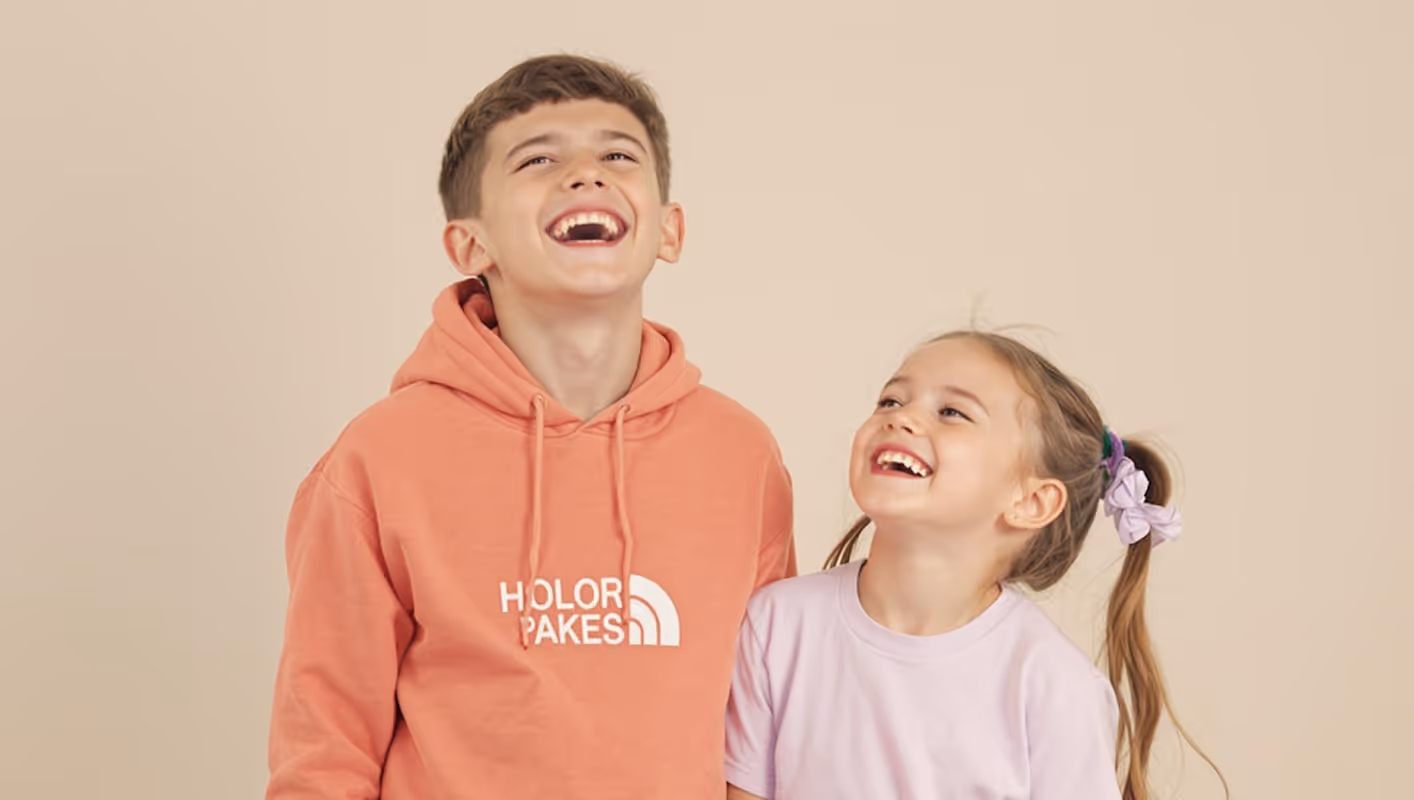 A boy in a peach hoodie and a girl in a lavender shirt laughing joyfully against a beige background.