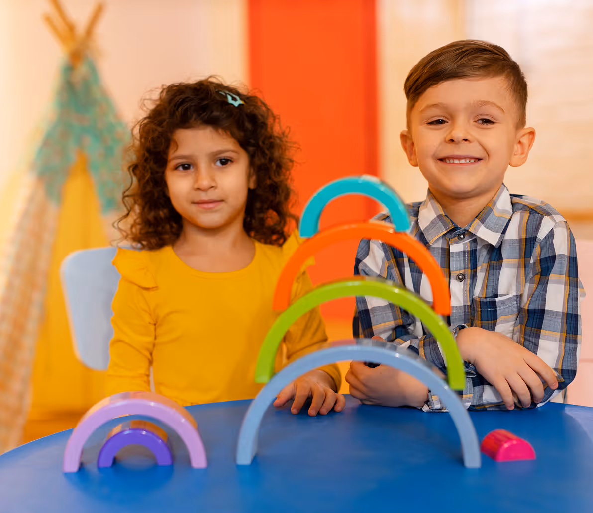Two children sitting at a blue table playing with colorful wooden rainbow stacker toys in a bright room.