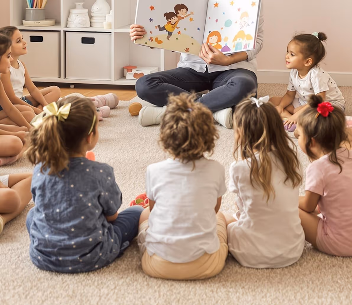 Group of young children sitting in a circle on a carpet listening to an adult reading a colorful storybook.