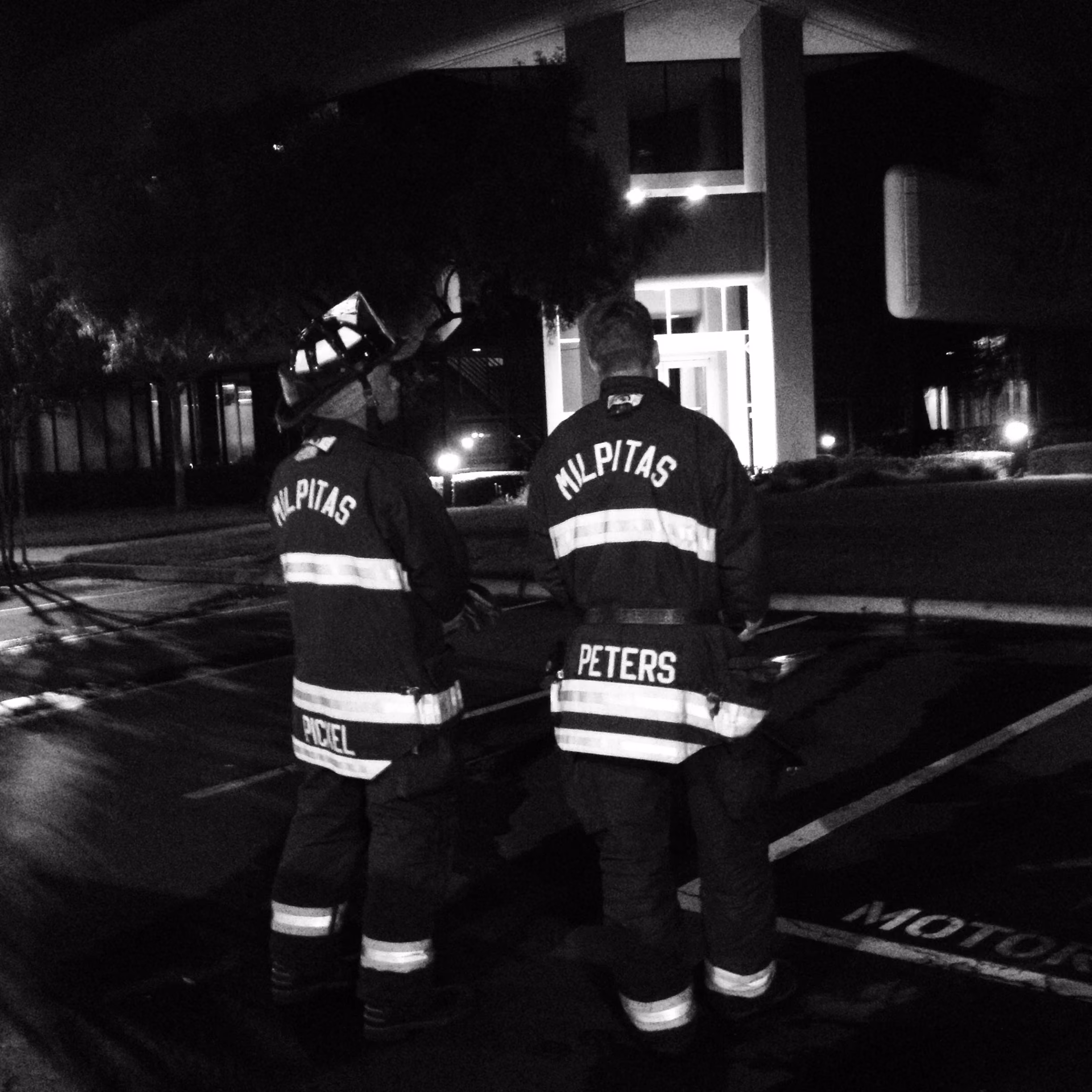 Two firefighters in Milpitas gear standing outside a building at night.