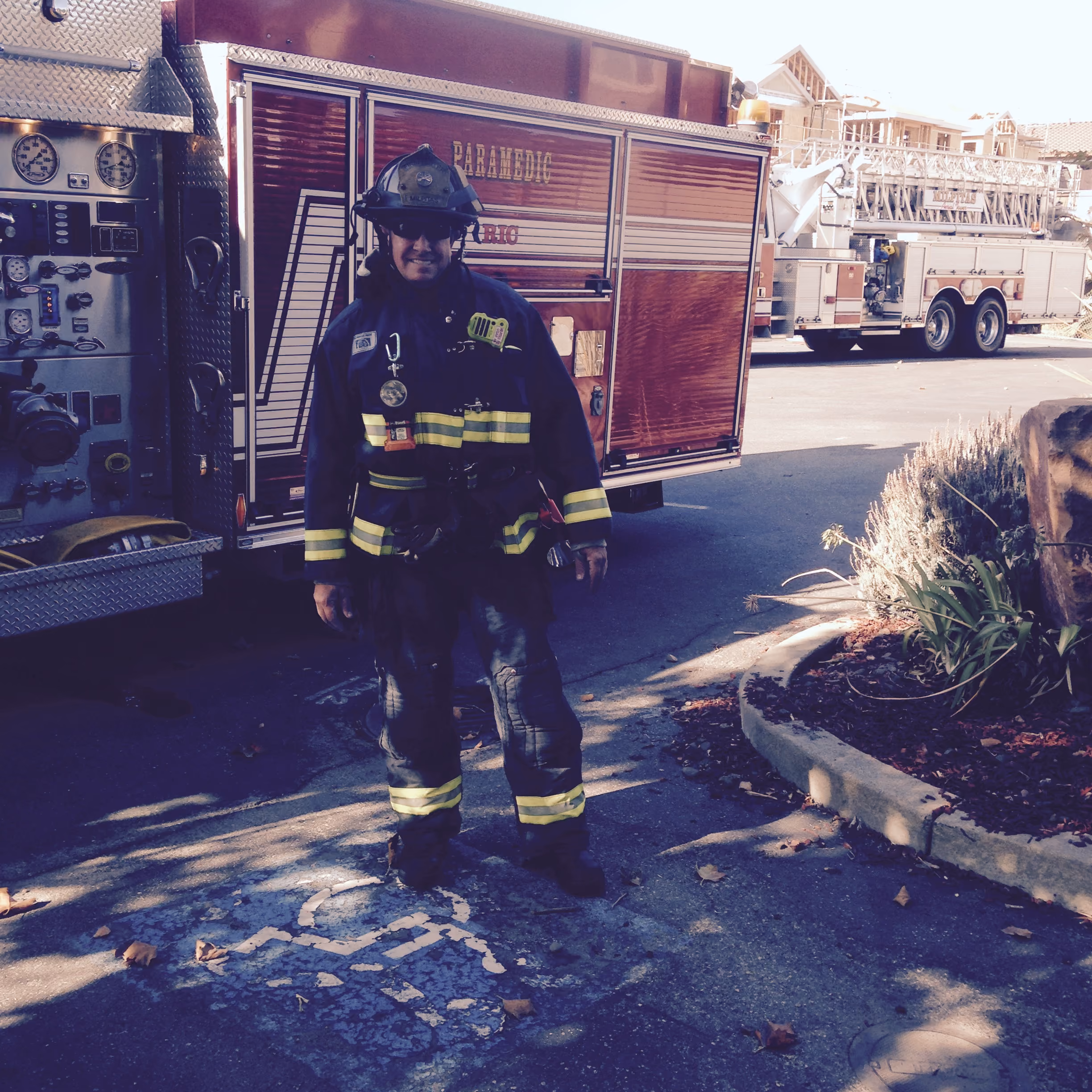 Firefighter in uniform standing in front of a parked fire truck on a sunny day.