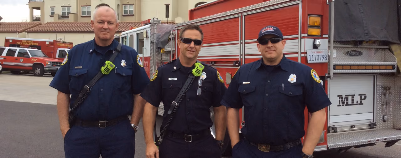 Three firefighters in navy uniforms standing in front of a red fire truck outside a building.