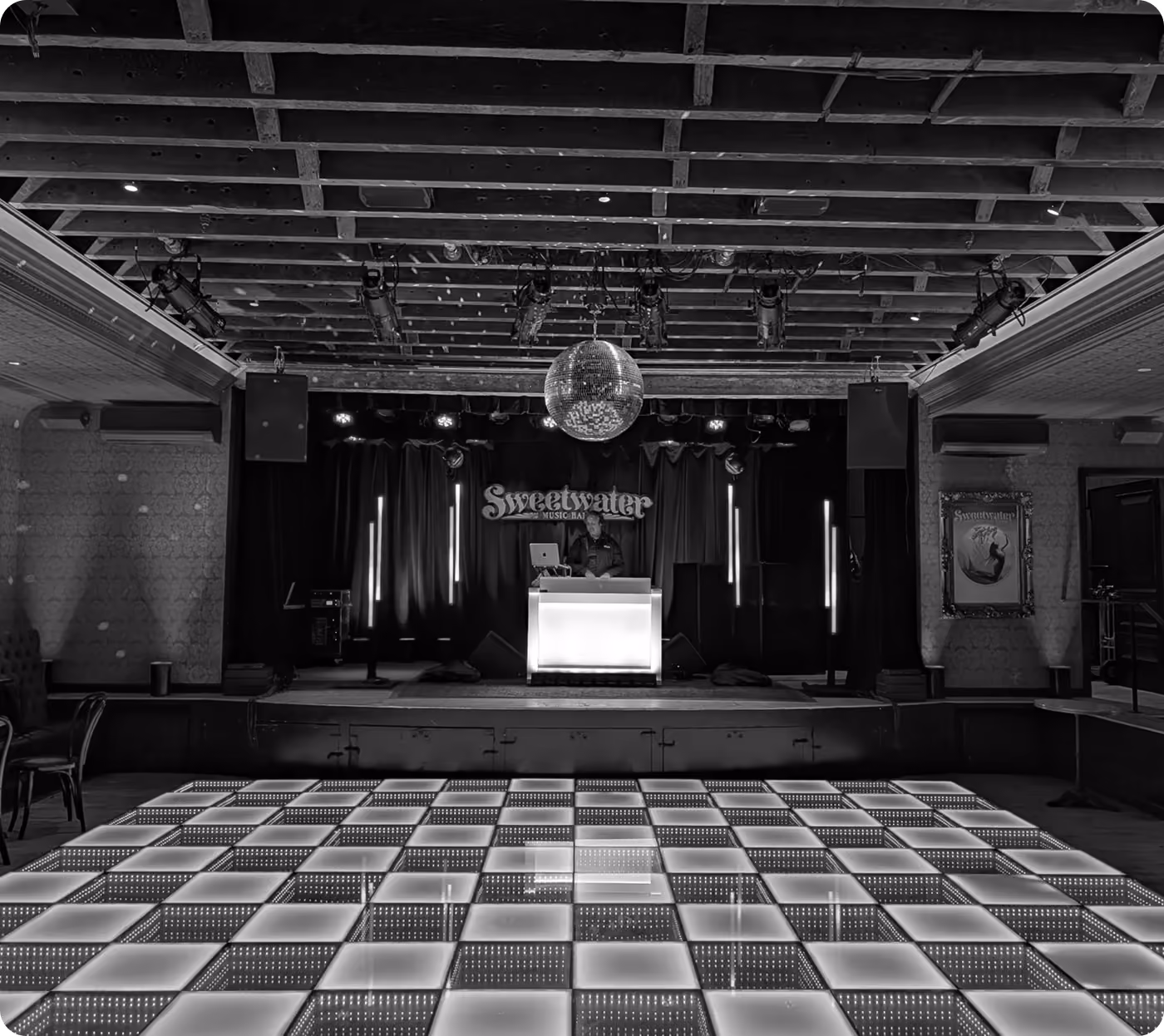 Black-and-white photo of an empty dance floor with a checkered floor, mirror ball and DJ booth