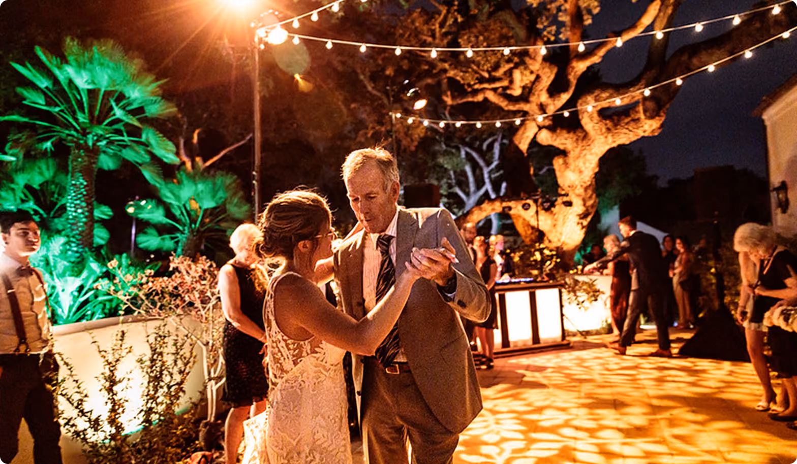 Couple dancing outdoors at night under string lights and an oak tree lit with green uplighting